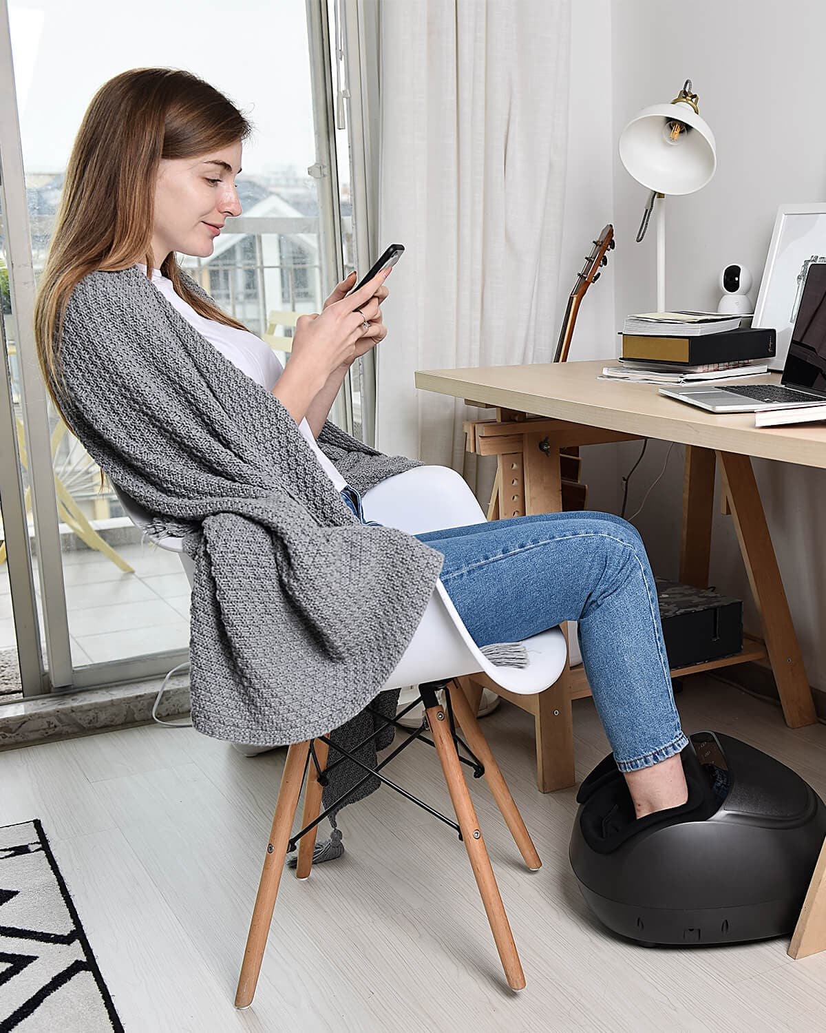 A woman sits in a modern white chair using her smartphone, draped in a gray cardigan and blue jeans. She is in a well-lit room with a desk, computer, and guitar while enjoying the Renpho EU Shiatsu Foot Massager Premium.