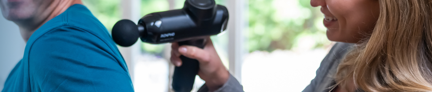 A woman with long hair smiles while using a black massage gun on the shoulder of a person wearing a blue shirt. The background shows a blurred window with greenery outside.