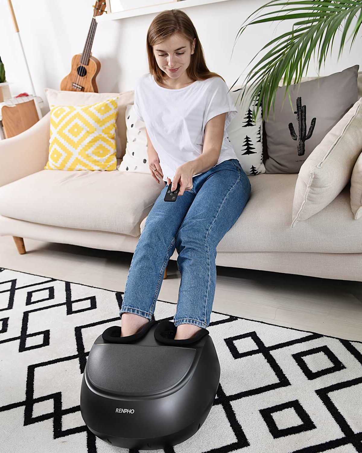 A young woman sitting on a cream couch using a black Renpho EU Shiatsu Foot Massager Premium in a cozy living room, with a guitar and potted plants in the background. She's dressed casually in a white t-shirt.