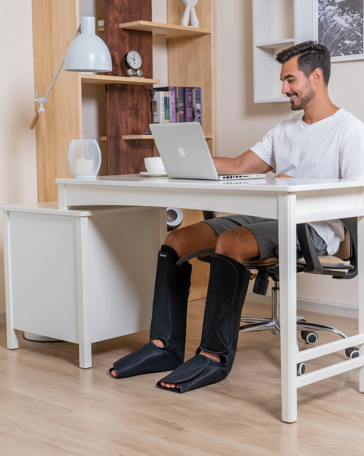 A man sits at a white desk on an office chair, using a laptop. He is wearing a white T-shirt and gray shorts. His legs are covered with Renpho EU Aeria Ultimate 2 Full Leg Massagers, extending from his feet to above his knees. The workspace includes shelves, books, a clock, and a lamp in the background.