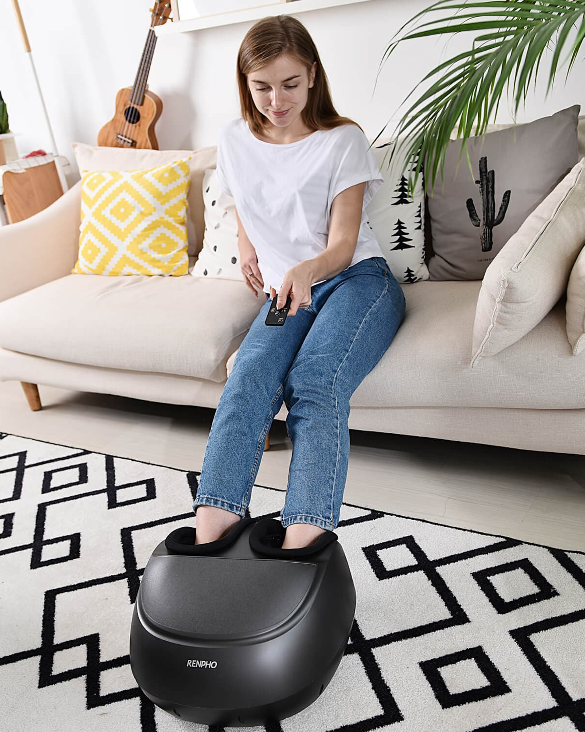 A woman in a white shirt and blue jeans uses a remote control to operate a Renpho EU Shiatsu Foot Massager Premium, relaxing her body with its deep kneading foot massage. She is sitting on a beige couch with patterned cushions, including one in yellow and white. There is a black and white rug on the floor and a cactus decoration on the couch. A guitar is seen in the background.
