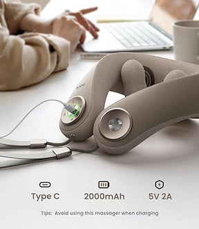 Close-up of an upgraded Renpho EU U-Neck Mini Neck Shoulder Massager on a white table, with charging cable attached to a Type-C port. Visible green light indicates charging. A laptop and coffee mug are in the blurred background. Illustration shows “2000mAh” battery capacity and “5V 2A” power rating. Note: Avoid using this massager when charging.